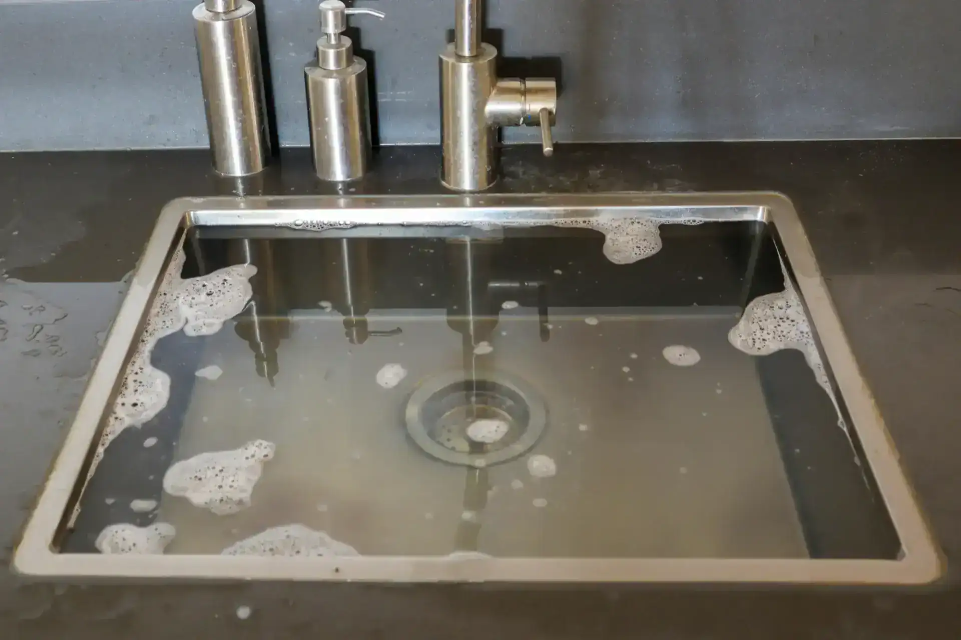 A stainless steel kitchen sink in FL is filled with soapy water, bubbles floating on the surface. Metal soap dispensers and a faucet sit on the countertop&mdash;perfect for a homeowner looking for a plumber Pasco County trusts.