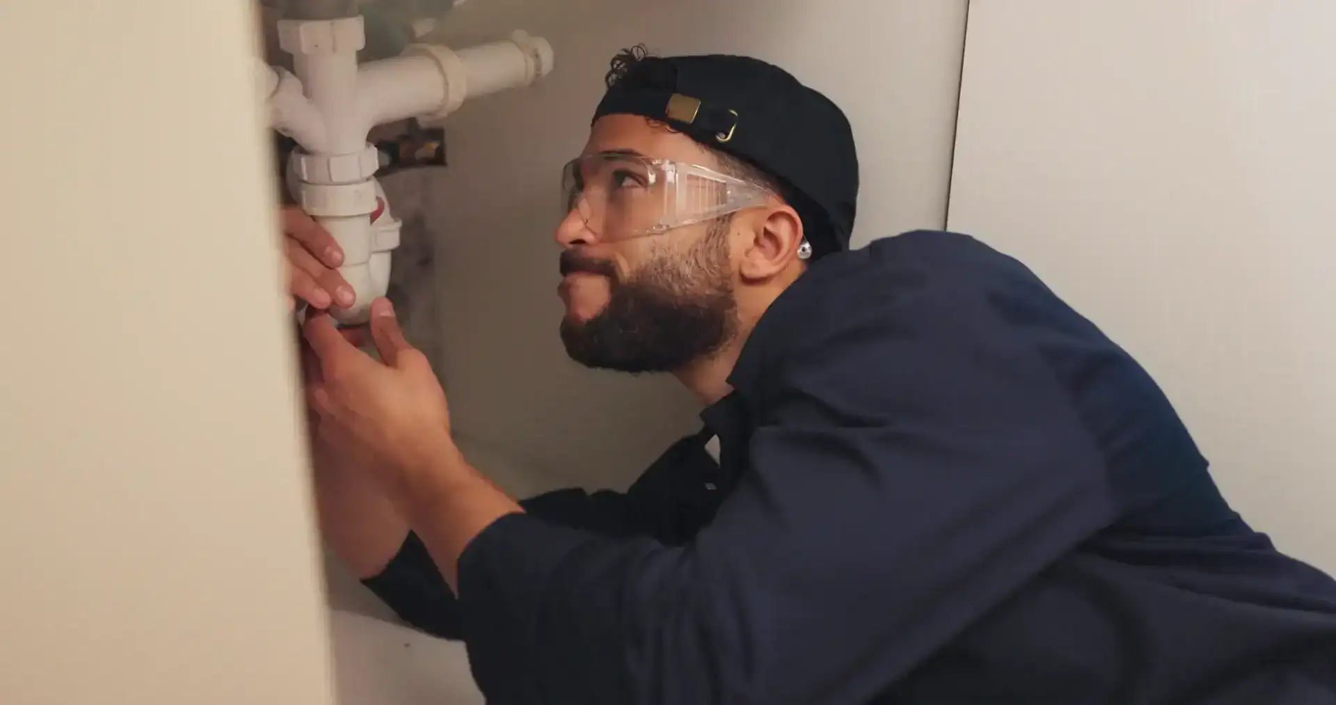 A man wearing safety goggles and a backwards cap repairs plumbing under a sink in FL, using his hands to examine the pipes—just like an expert plumber Pasco County relies on.