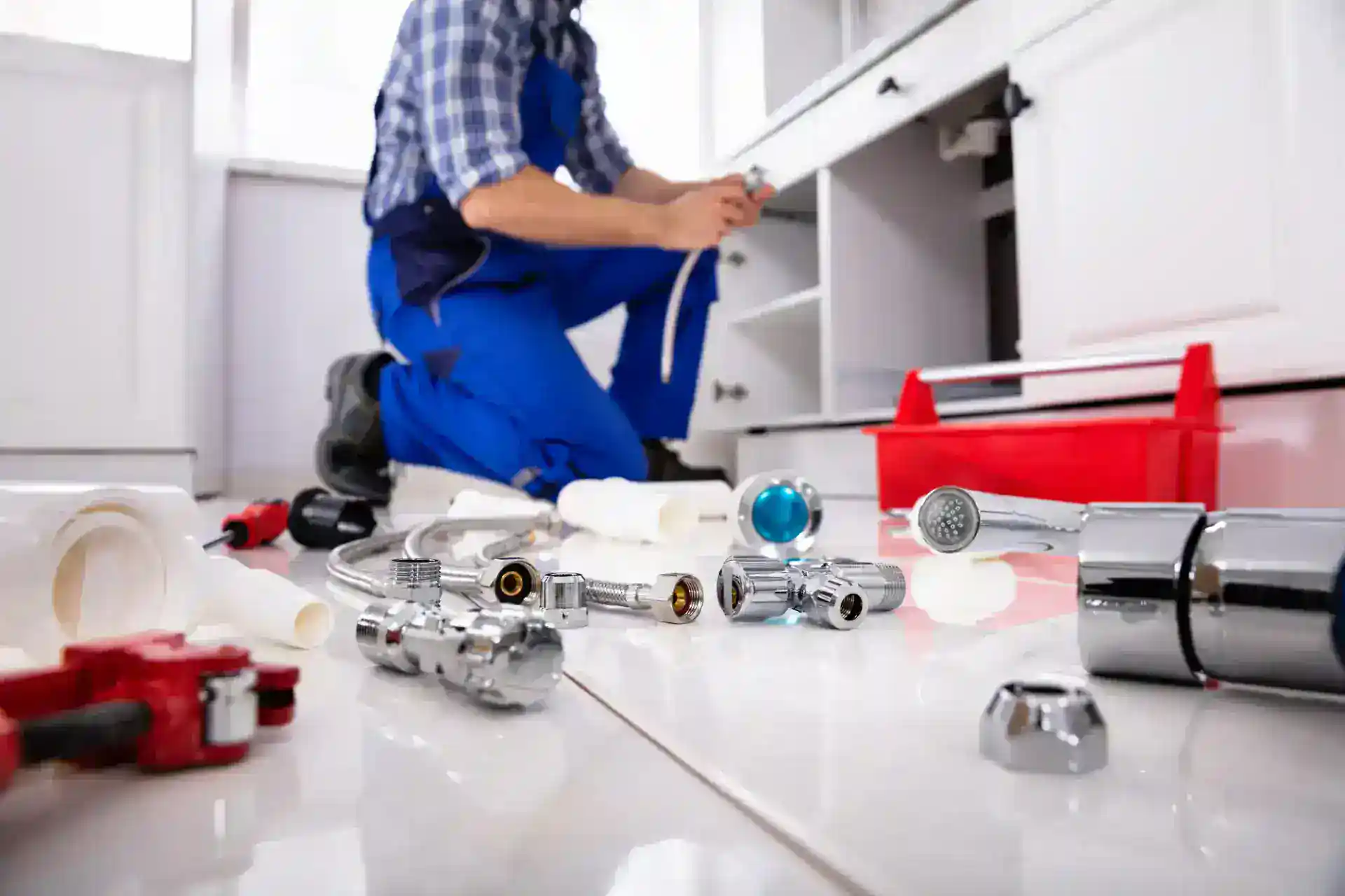 A plumber in blue overalls kneels on a kitchen floor, working under a sink with various plumbing tools and parts scattered on the white tile floor, next to a red toolbox.